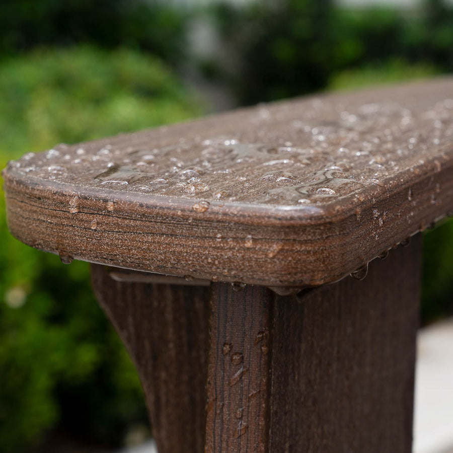 Close-up of a rain-soaked bench showing droplets of water on the wood surface. The background is blurred with lush green foliage, highlighting the wet texture and detail of the bench.