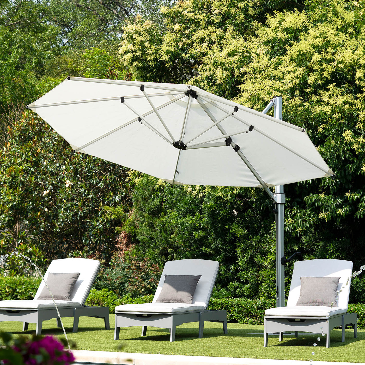 Four gray lounge chairs with gray cushions are arranged on a lawn near a pool. A large, white cantilever umbrella provides shade over two chairs. The background features lush green trees and bushes under a clear sky.
