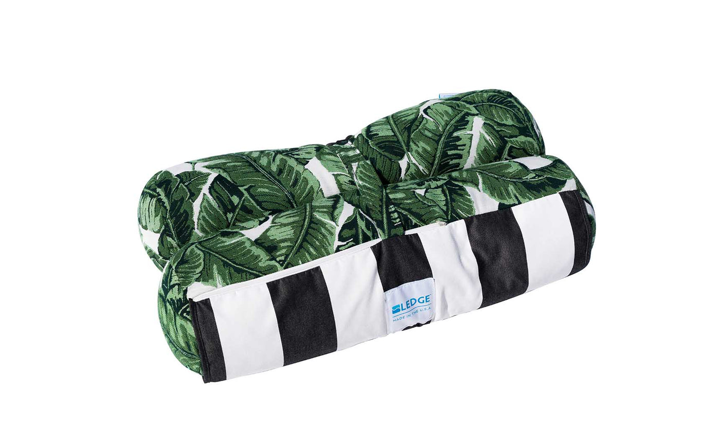 This decorative pillow showcases green banana leaves on one side and black and white stripes on the other. Made from marine-grade fabric, its ideal for outdoor use. The blue Ledge Lounger label is visible at its rectangular edge, perfect for your Laze Hammock or patio chair.