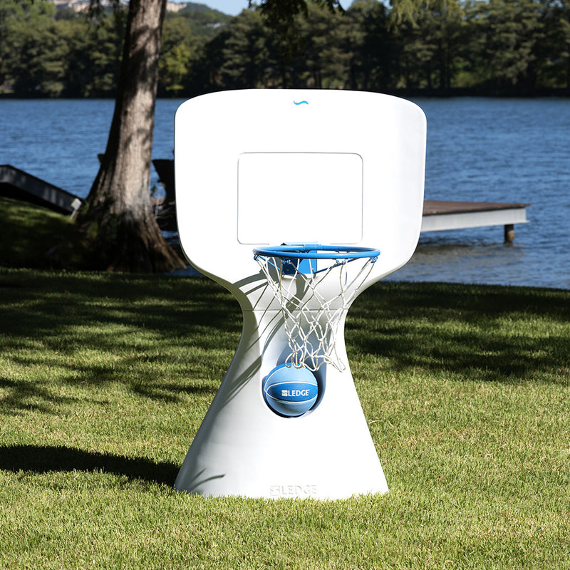 A sleek, modern white basketball hoop with a blue net stands on a grassy area by a lake. A wooden dock and trees are visible in the background, with sunlight casting shadows on the ground.