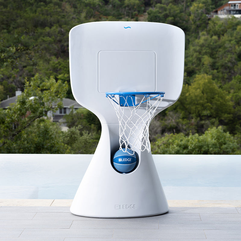 Modern white basketball hoop with a blue net, positioned on a poolside with a scenic view of lush green trees and hills in the background. The hoop is placed on a stone-tiled pool deck.