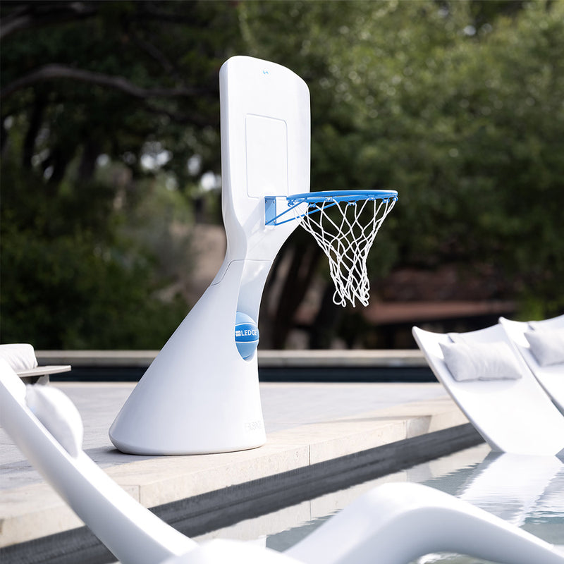 A sleek, modern white basketball hoop with a blue rim stands beside a pool. It features a smooth, curved base design. In the foreground and background are white lounge chairs, and a lush green landscape is visible in the distance.