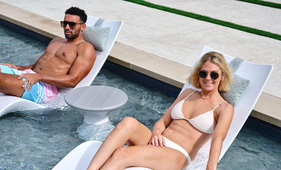 A man in colorful shorts and a woman in a white bikini, both wearing sunglasses, relax on white pool loungers partially submerged in water. Beside them is the sleek Autograph Standard Side Table by Ledge Lounger, complementing their sunny escape.
