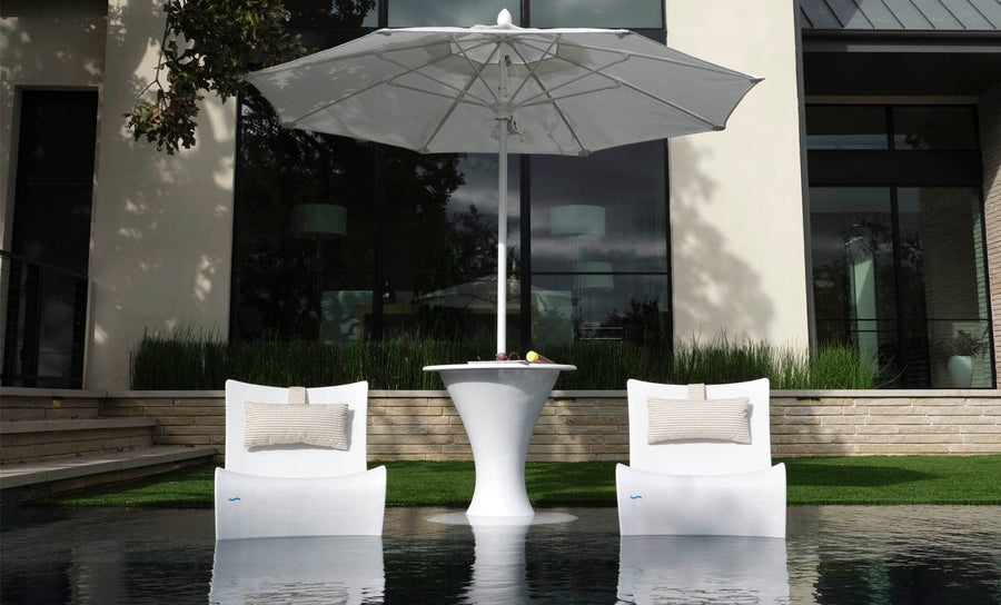 Two modern white lounge chairs in water face a Ledge Lounger Autograph Umbrella Stand Ice Bin and tall outdoor table. A drink and fruit are on the table, set outside a contemporary building with large windows and lush greenery.