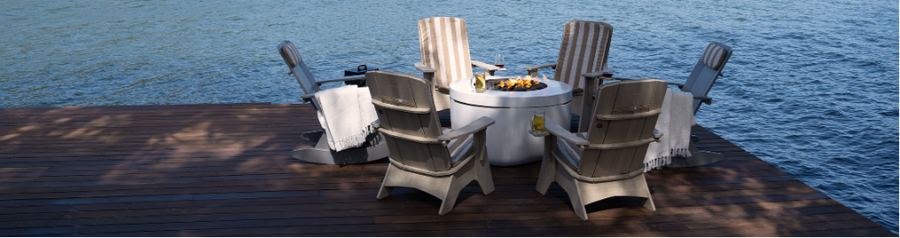 Five Adirondack chairs with blankets are arranged around a fire pit on a wooden dock overlooking a calm body of water. Drinks rest on the armrests, creating a cozy outdoor setting.