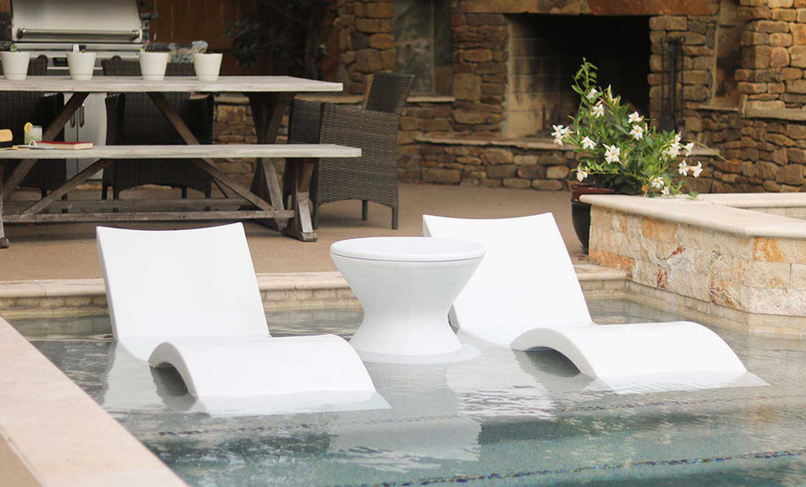 A pair of Ledge Loungers Outdoor Oasis In-Pool Furniture Bundles, featuring deep chaise loungers in white, are partially submerged in a shallow pool. An ice bin side table sits nearby against a stone wall with plants and a wooden dining table set in the background.