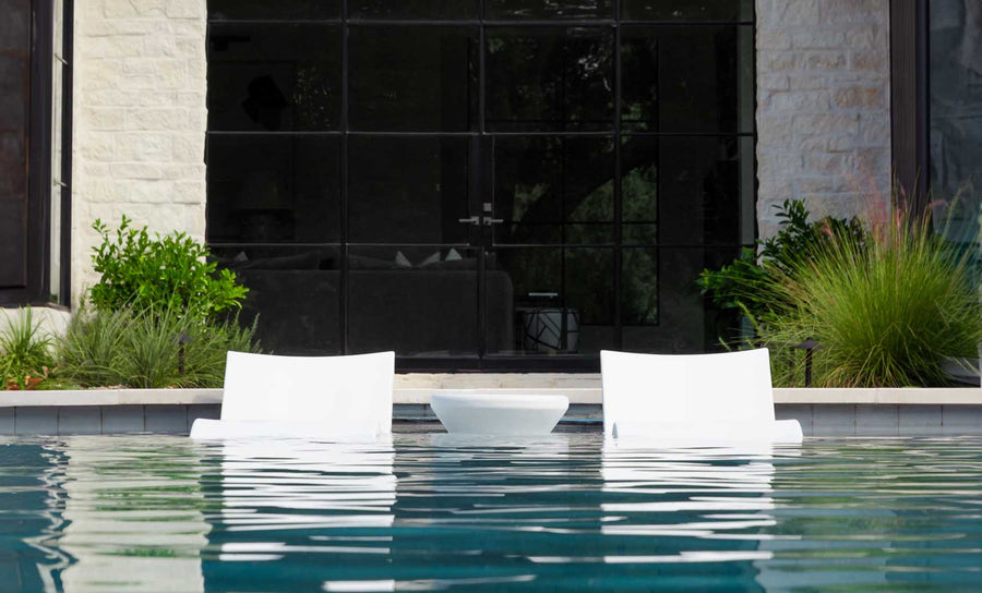 Two white Outdoor Oasis In-Pool Furniture Bundle loungers by Ledge Lounger sit partially submerged in the pool, facing a modern building with large glass windows. The clear water reflects the loungers, offering an exquisite escape amidst lush greenery.