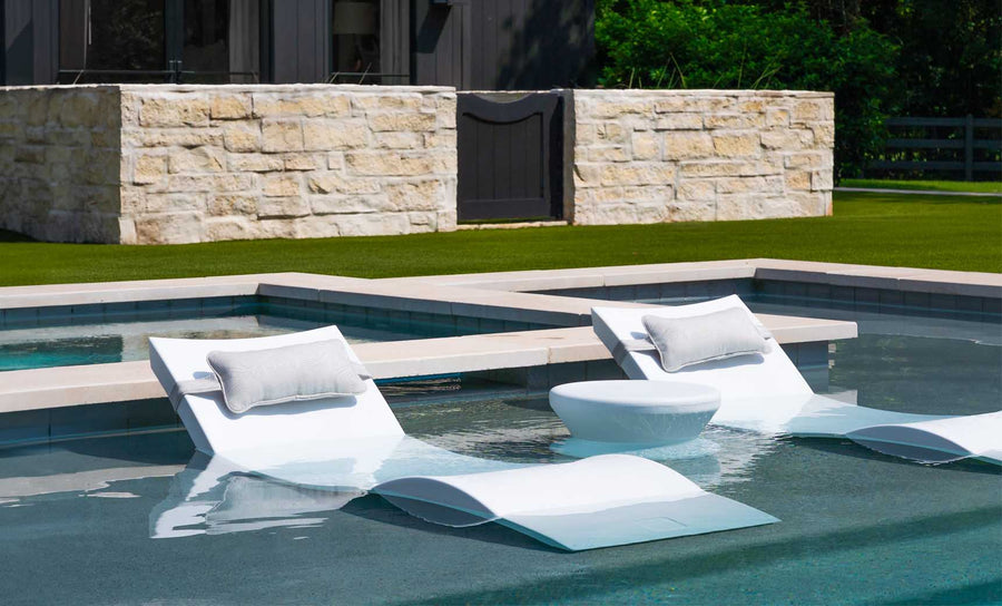 Two Ledge Lounger white in-pool chaise loungers with pillows, accompanied by a small round table, are semi-submerged in a shallow pool. A stone wall and grassy lawn add to the scenes tranquility. Enjoy the sunny escape with the Outdoor Paradise In-Pool Furniture Bundle.