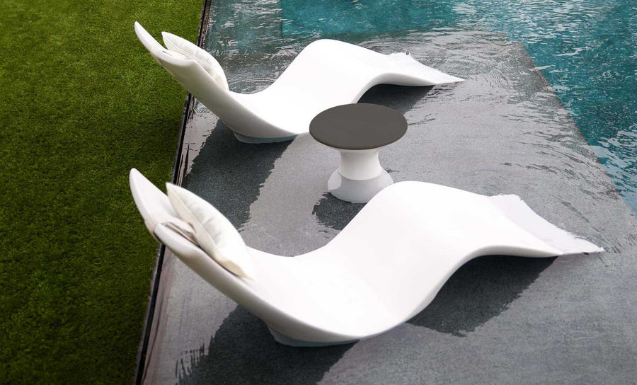The Resort Luxury Outdoor Paradise In-Pool Furniture Bundle from Ledge Lounger features two wave-shaped, modern white chaise loungers partially submerged in a pool beside a small, black-topped round table, creating a serene and sunny poolside retreat where the pool meets the grassy lawn.