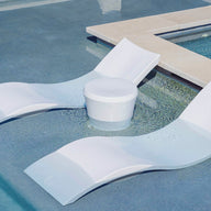 A pair of white, curved lounge chairs and a Ledge Lounger Affinity Side Table sit partially submerged in the shallow pool, seamlessly blending into the serene setting as tranquil outdoor accents with water lapping gently over them.