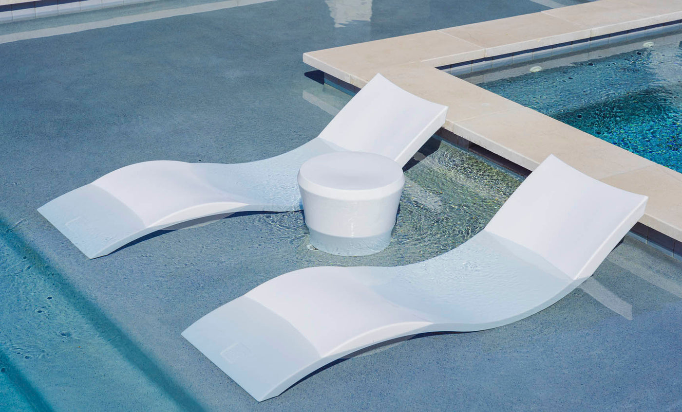 A pair of white, curved lounge chairs and a Ledge Lounger Affinity Side Table sit partially submerged in the shallow pool, seamlessly blending into the serene setting as tranquil outdoor accents with water lapping gently over them.