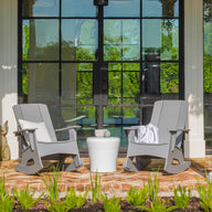 Two gray rocking chairs on a brick porch have a Ledge Lounger Affinity Side Table between them. Reflections of the garden greenery in a glass door enhance the scene, while a white column and potted plants frame this inviting outdoor area, highlighting chic accessory potential.