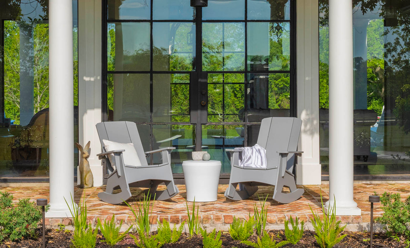 Two gray rocking chairs on a brick porch have a Ledge Lounger Affinity Side Table between them. Reflections of the garden greenery in a glass door enhance the scene, while a white column and potted plants frame this inviting outdoor area, highlighting chic accessory potential.