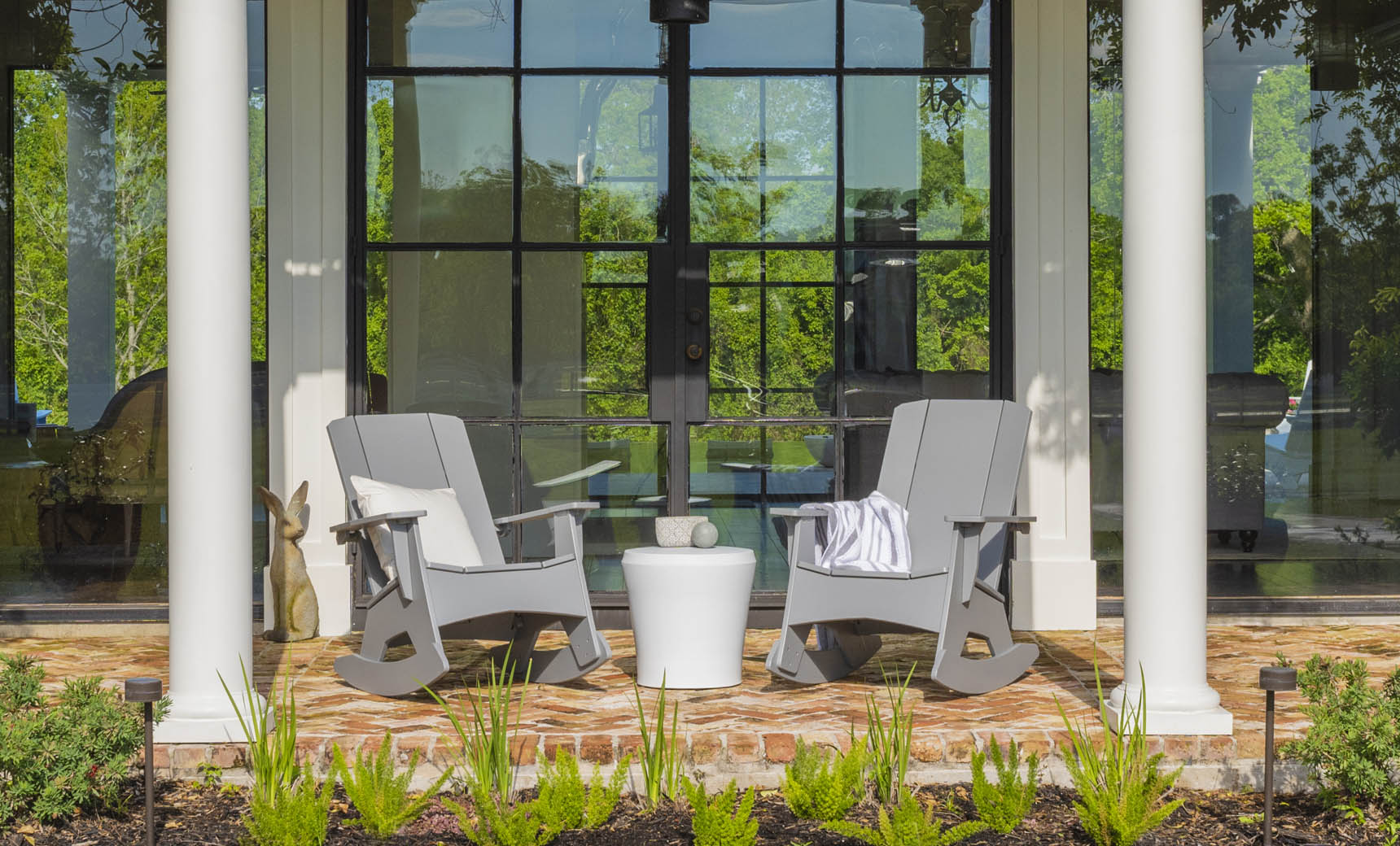 Two gray rocking chairs on a brick porch have a Ledge Lounger Affinity Side Table between them. Reflections of the garden greenery in a glass door enhance the scene, while a white column and potted plants frame this inviting outdoor area, highlighting chic accessory potential.