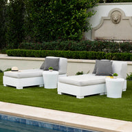 Two white lounge chairs with gray cushions and Ledge Lounger Affinity Side Tables are placed on a green lawn by the pool. A beige sun hat is on one chair. The setting includes a stone wall with a carved lions head and lush greenery, forming an inviting oasis for relaxation.