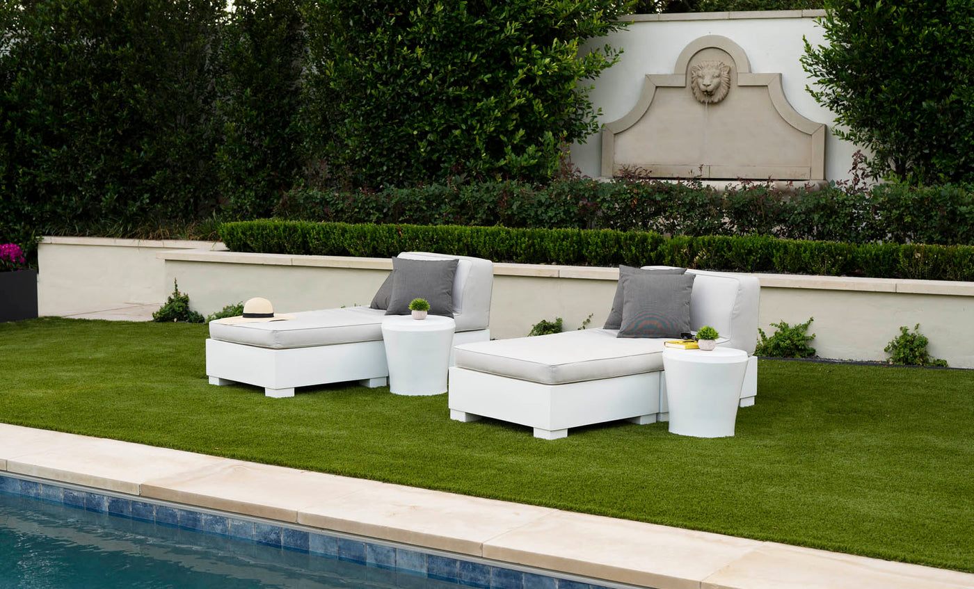 Two white lounge chairs with gray cushions and Ledge Lounger Affinity Side Tables are placed on a green lawn by the pool. A beige sun hat is on one chair. The setting includes a stone wall with a carved lions head and lush greenery, forming an inviting oasis for relaxation.