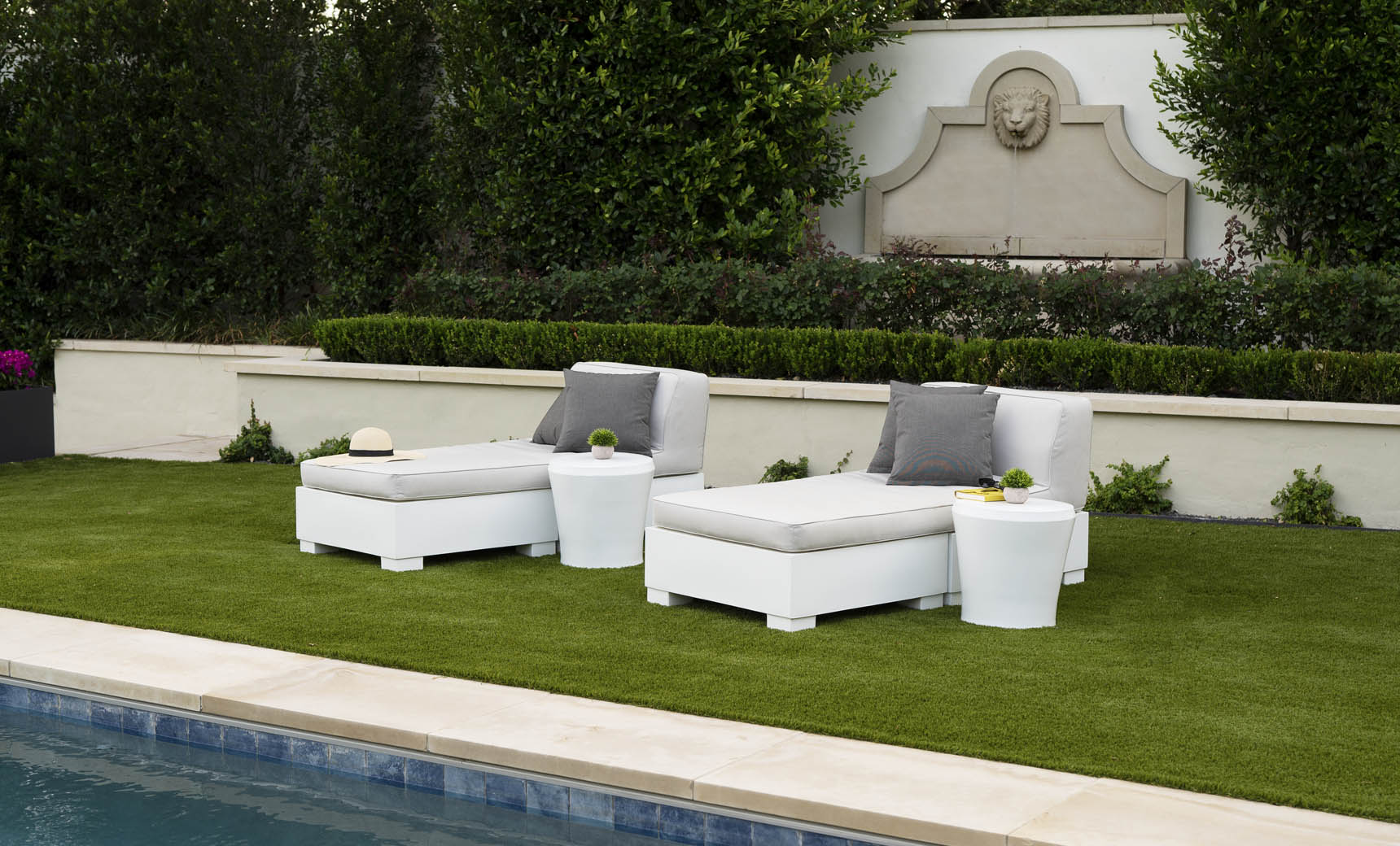 Two white lounge chairs with gray cushions and Ledge Lounger Affinity Side Tables are placed on a green lawn by the pool. A beige sun hat is on one chair. The setting includes a stone wall with a carved lions head and lush greenery, forming an inviting oasis for relaxation.