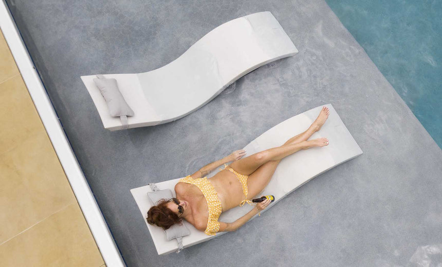 A person in a yellow bikini relaxes on a Signature Chaise from Ledge Lounger next to a pool, drink in hand. Beside them is another empty lounger with pillows. The scene perfectly captures the relaxing essence of Ledge Loungers Outdoor Escape In-Pool Furniture Bundle in the sunny poolside setting.