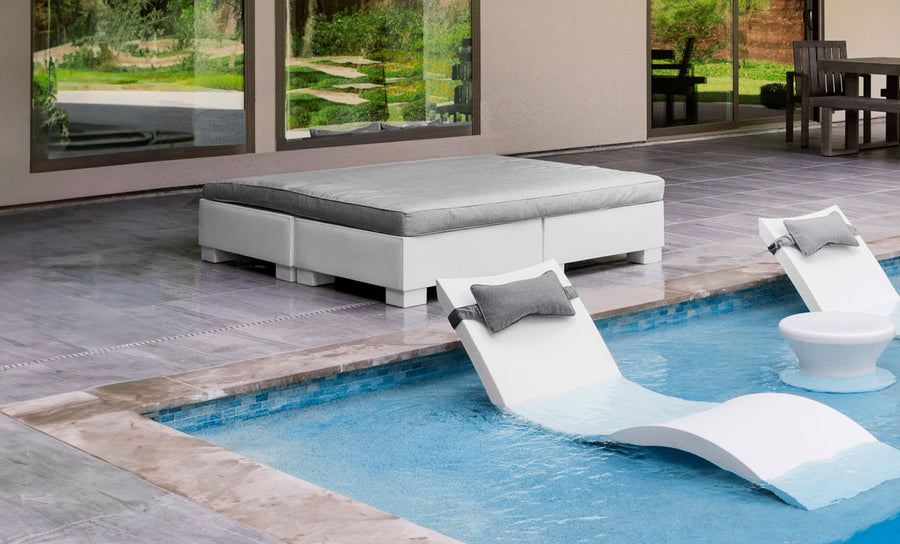 An outdoor area showcases an Affinity Square Sunbed with a Flat Cushion from Ledge Lounger, accompanied by two white lounge chairs partially submerged in the pool. A cushioned square seating area sits nearby on the gray-tiled patio, while large windows reflect greenery.