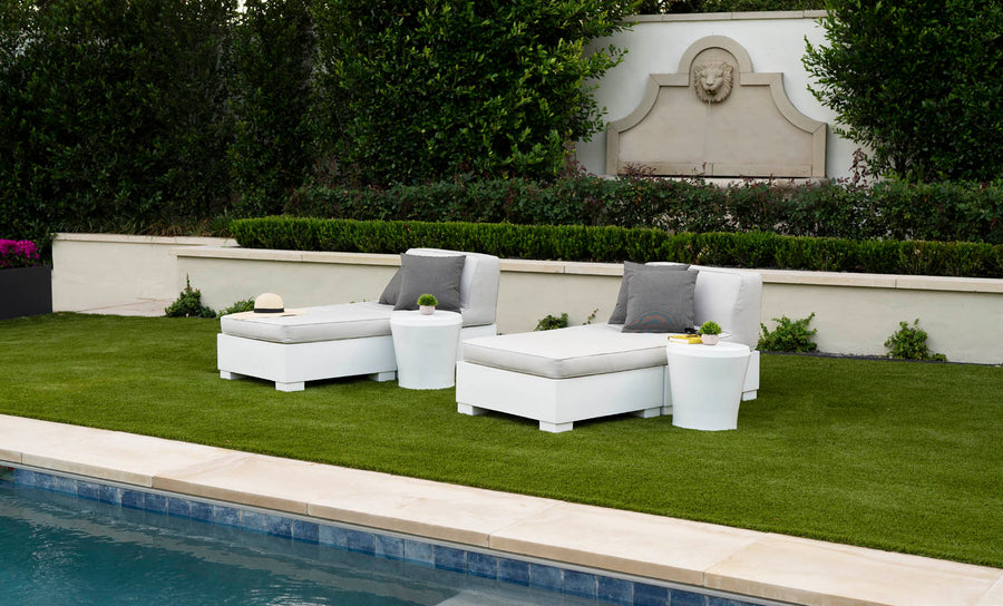Two white Ledge Lounger Affinity 2 Piece Sun Chairs with cushions and small tables, made from UV20 rated resin, are on a grassy spot by the pool. A hat and potted plants decorate the tables. In the background, a stone wall with greenery features a decorative lions head fountain.