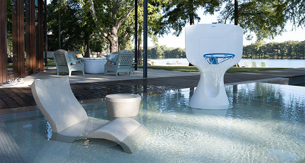 A serene outdoor pool scene with a contemporary lounge chair and small table set partially in the water. A white basketball hoop is also in the pool area. In the background, theres a seating area with chairs, trees, and a view of the lake.