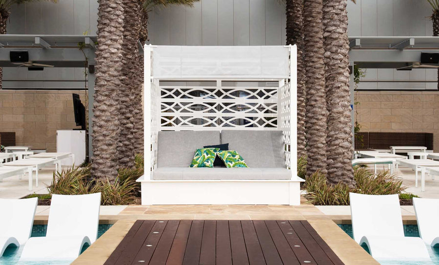 Outdoor lounge area with a customizable Ledge Lounger Shift Daybed in white, nestled between palm trees. Patterned cushions adorn the daybed by the pool. White lounge chairs decorate the deck, framed by modern architecture and made from durable outdoor materials.
