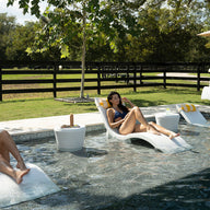 Two people are lounging on chairs partially submerged in a pool. They are relaxing under a tree on a sunny day, with drinks on small tables beside them. A fence and grassy area are visible in the background.