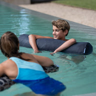 A boy and girl float in a pool using Laze Doodle cushions from Ledge Lounger. The boy smiles at the girl, who faces away from the camera. The outdoor pool is surrounded by grass and a gravel path.