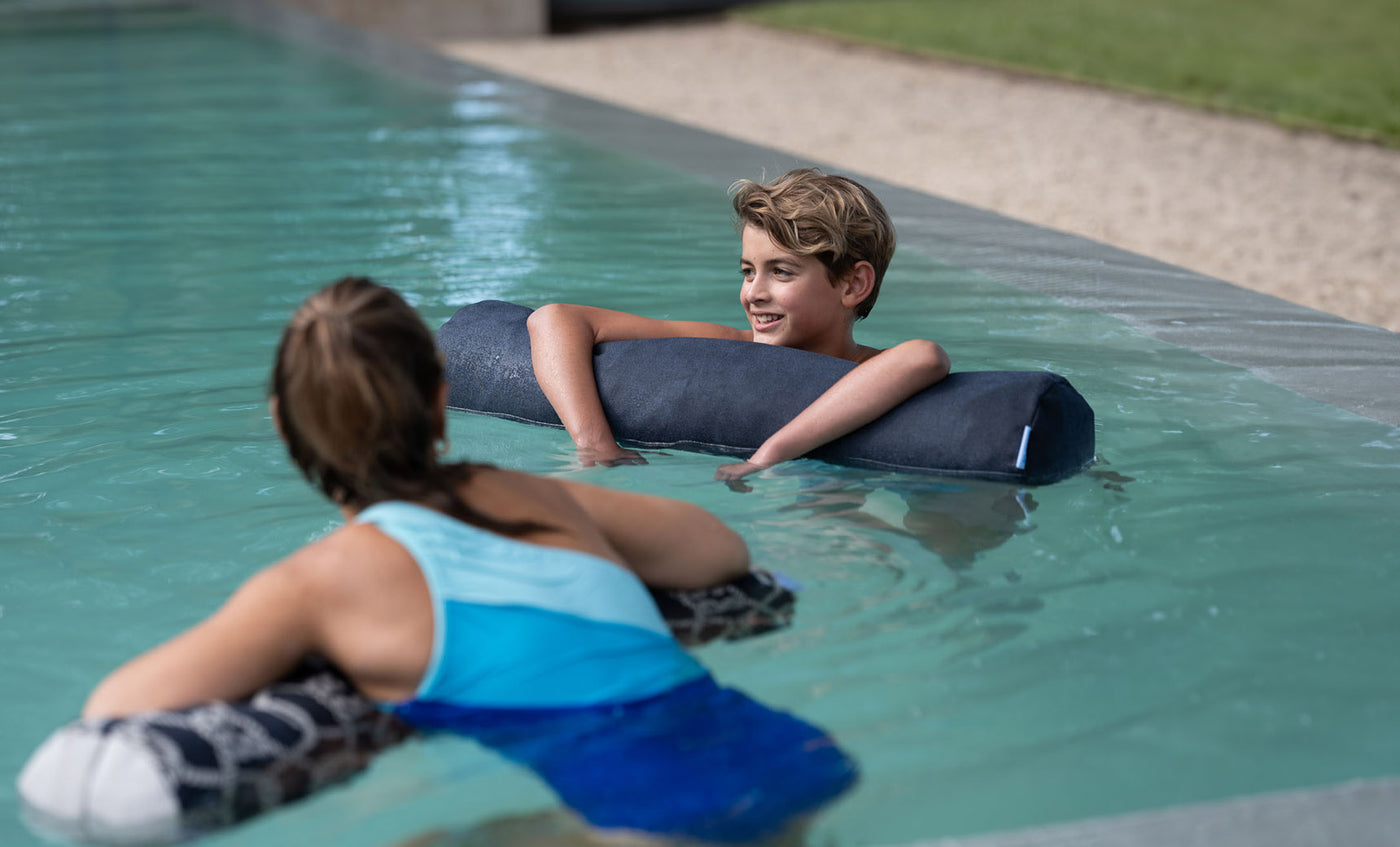 A boy and girl float in a pool using Laze Doodle cushions from Ledge Lounger. The boy smiles at the girl, who faces away from the camera. The outdoor pool is surrounded by grass and a gravel path.