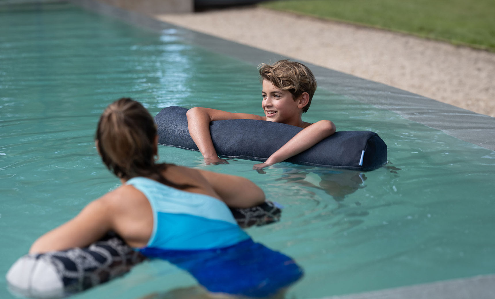 A boy and girl float in a pool using Laze Doodle cushions from Ledge Lounger. The boy smiles at the girl, who faces away from the camera. The outdoor pool is surrounded by grass and a gravel path.