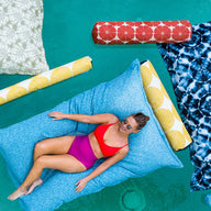 Wearing a red and pink swimsuit, someone relaxes on a large blue Laze Doodle float by Ledge Lounger in a pool. They are surrounded by colorful patterned floats featuring yellow, red, and blue designs in the clear turquoise water.