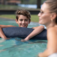 A young boy smiles while leaning on a Laze Doodle by Ledge Lounger in the pool. A woman with sunglasses is partially turned towards him, both looking relaxed and happy with trees and grass in the background.