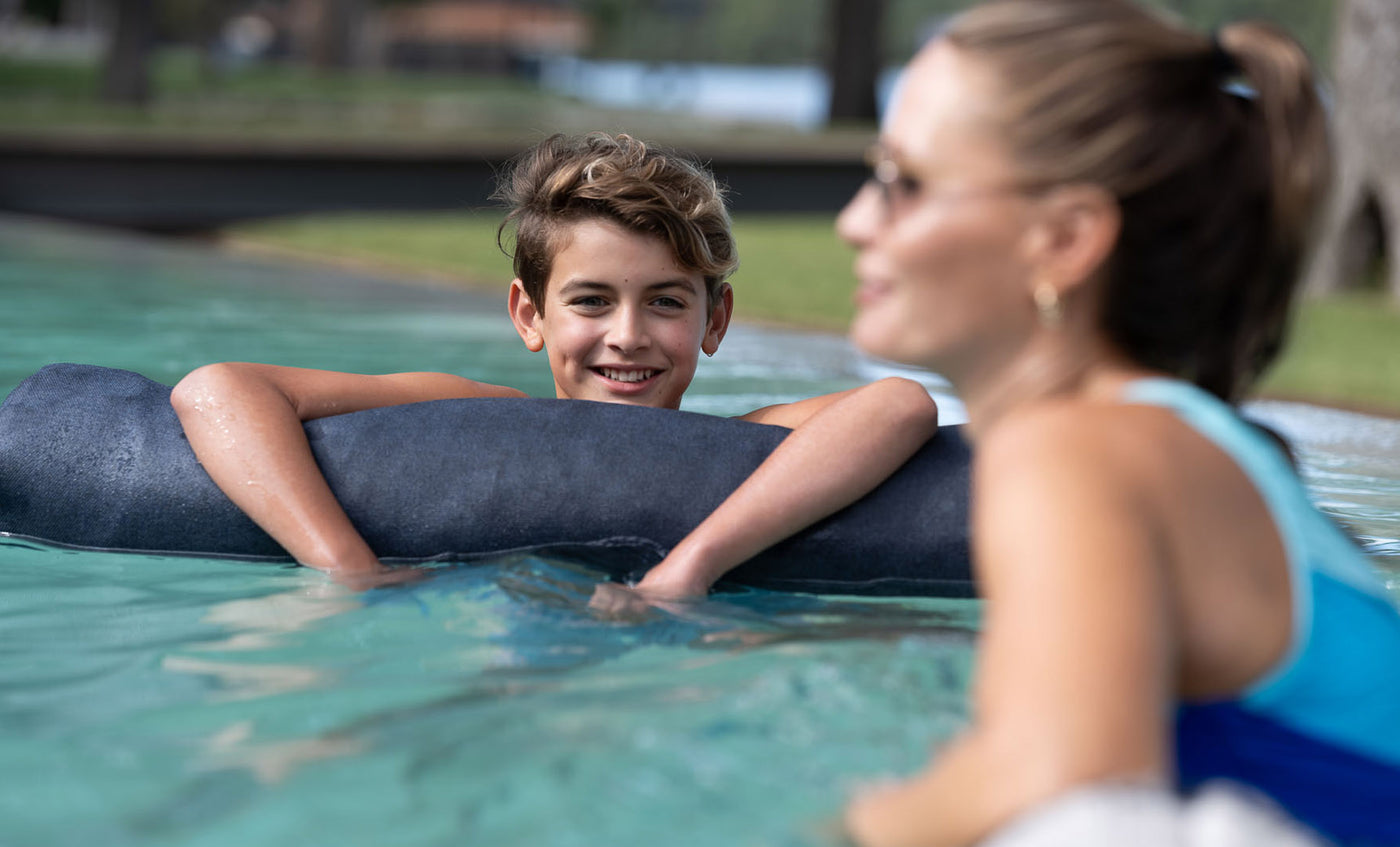 A young boy smiles while leaning on a Laze Doodle by Ledge Lounger in the pool. A woman with sunglasses is partially turned towards him, both looking relaxed and happy with trees and grass in the background.
