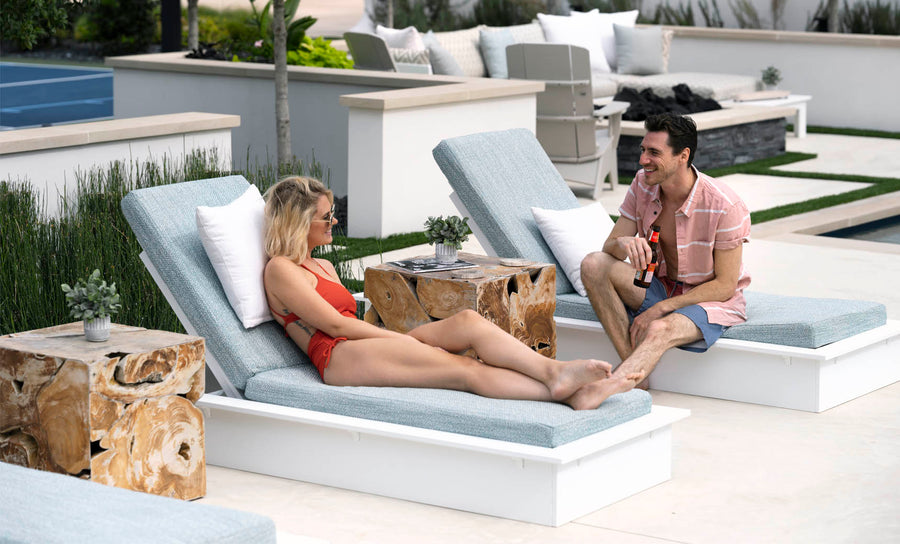 Reclining on the Ledge Lounger Echo Chaise, a woman in a red swimsuit and sunglasses smiles at a man in a pink shirt nearby. They relax among potted plants, making the most of their modern outdoor setting.