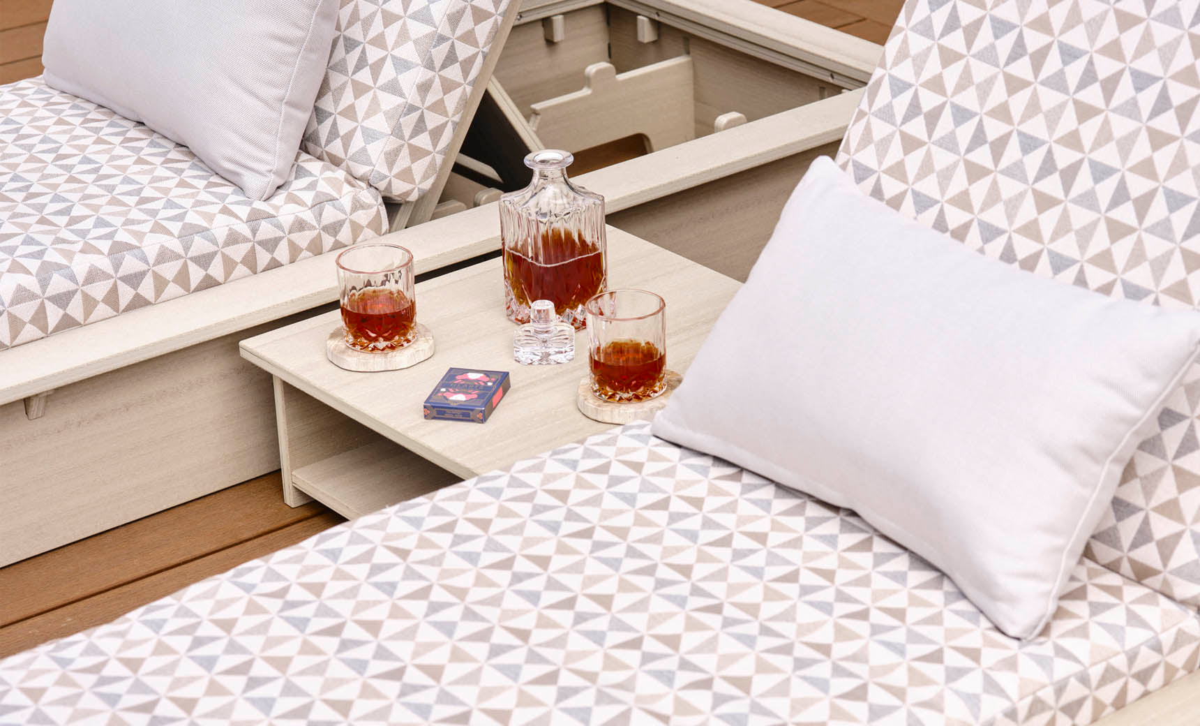 Two patterned lounge chairs with triangle designs, each topped with a white pillow. The Echo Side Table by Ledge Lounger holds a decanter, two glasses filled with amber liquid, and a deck of cards on a wooden deck.