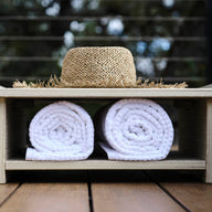 An Echo Side Table by Ledge Lounger sits with a straw hat on its smooth wooden shelf, positioned above two rolled white towels. The outdoor setting features blurred greenery in the background, creating a serene atmosphere.