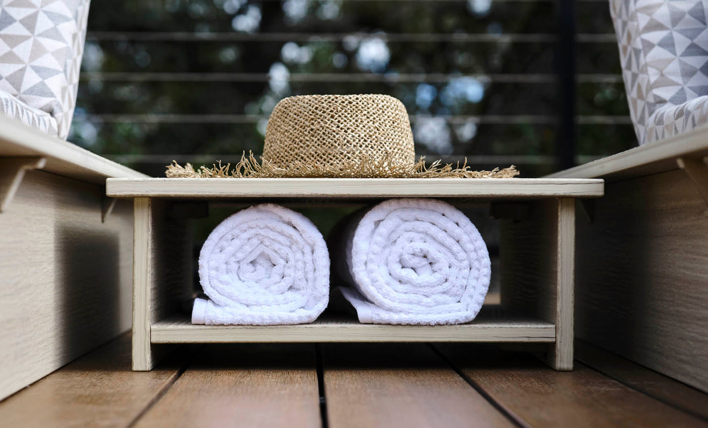 An Echo Side Table by Ledge Lounger sits with a straw hat on its smooth wooden shelf, positioned above two rolled white towels. The outdoor setting features blurred greenery in the background, creating a serene atmosphere.