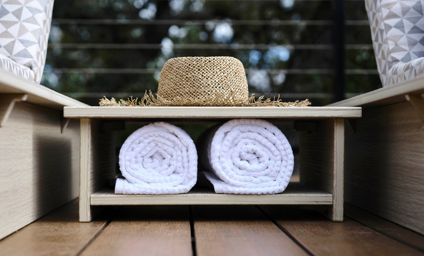 An Echo Side Table by Ledge Lounger sits with a straw hat on its smooth wooden shelf, positioned above two rolled white towels. The outdoor setting features blurred greenery in the background, creating a serene atmosphere.