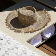 A straw hat sits on the Echo Side Table by Ledge Lounger, next to a lounge chair with a geometric-patterned cushion. Two rolled white towels are neatly placed in a compartment beneath the table.
