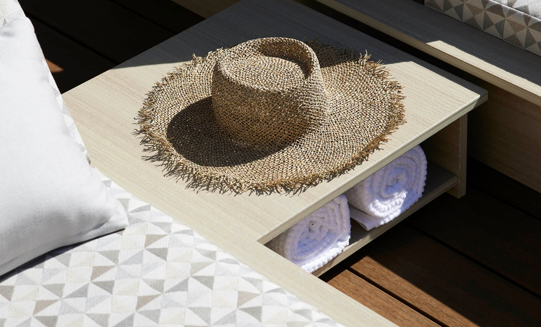 A straw hat sits on the Echo Side Table by Ledge Lounger, next to a lounge chair with a geometric-patterned cushion. Two rolled white towels are neatly placed in a compartment beneath the table.