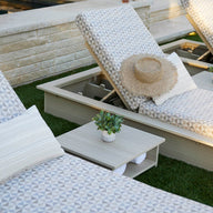 Two Ledge Lounger patterned poolside loungers with pillows sit on a lawn by the pool, one topped with a straw hat. Echo Side Tables - Final Sale, each adorned with small potted plants, are positioned between the loungers for added decor.
