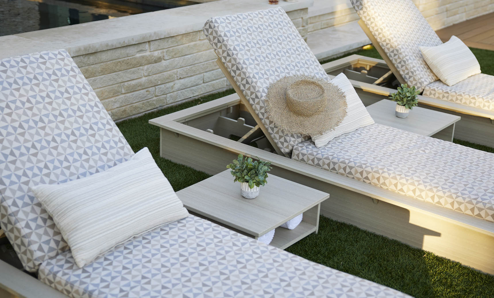 Two Ledge Lounger patterned poolside loungers with pillows sit on a lawn by the pool, one topped with a straw hat. Echo Side Tables - Final Sale, each adorned with small potted plants, are positioned between the loungers for added decor.