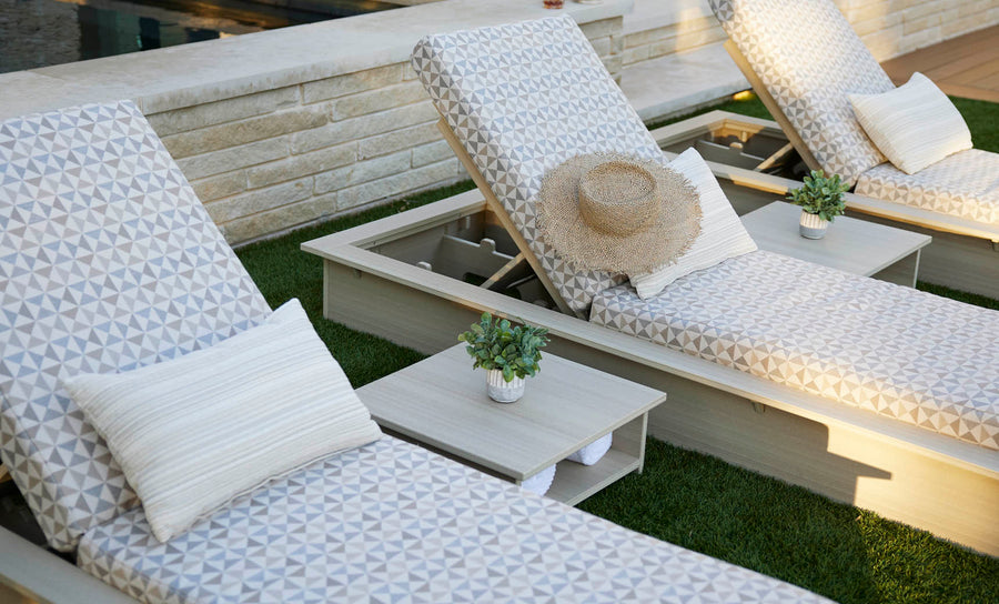 Two Ledge Lounger patterned poolside loungers with pillows sit on a lawn by the pool, one topped with a straw hat. Echo Side Tables - Final Sale, each adorned with small potted plants, are positioned between the loungers for added decor.