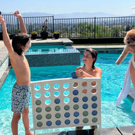 A woman in a swimsuit and sunhat holds a fluffy dog by the pool, while a young woman smiles from within it. Nearby, two children raise their arms in excitement beside Ledge Loungers Four In A Row: an oversized outdoor game with large colored chips, set against a bright sky and mountains.