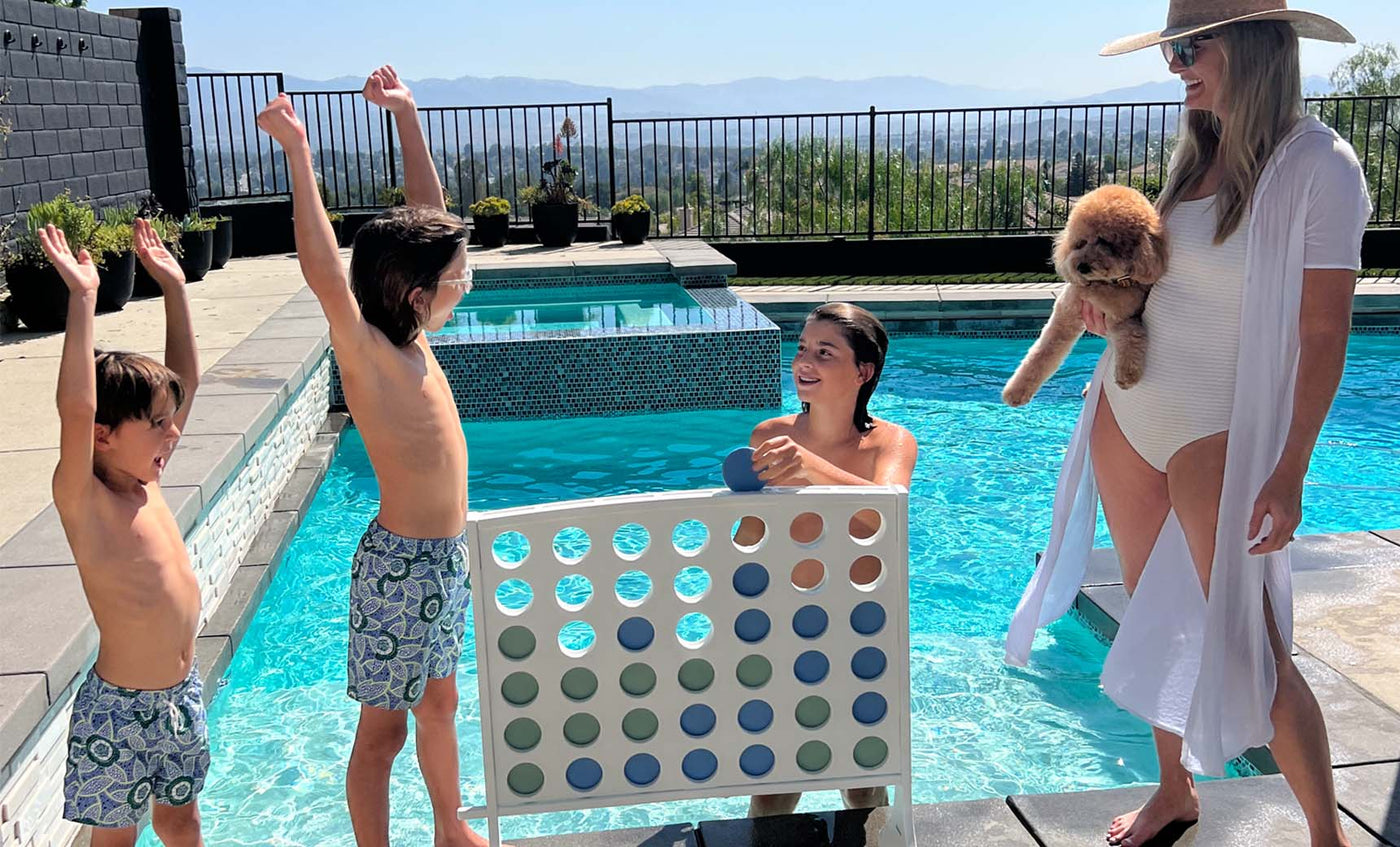 A woman in a swimsuit and sunhat holds a fluffy dog by the pool, while a young woman smiles from within it. Nearby, two children raise their arms in excitement beside Ledge Loungers Four In A Row: an oversized outdoor game with large colored chips, set against a bright sky and mountains.