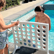 Two boys in swim trunks enjoy oversized outdoor fun at the pools edge, playing Ledge Loungers Four In A Row. One places a large colored chip while the other looks on, surrounded by sunlit water and tiled edges.