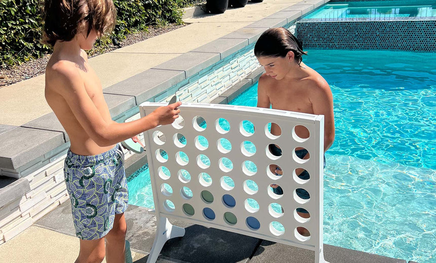 Two boys in swim trunks enjoy oversized outdoor fun at the pools edge, playing Ledge Loungers Four In A Row. One places a large colored chip while the other looks on, surrounded by sunlit water and tiled edges.