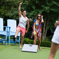 Two people enjoy a lively game of Ledge Lounger Cornhole outdoors. One person celebrates while the other smiles with a drink. Nearby, two others stand by the cornhole set, surrounded by large blue chairs and lush greenery in the background.