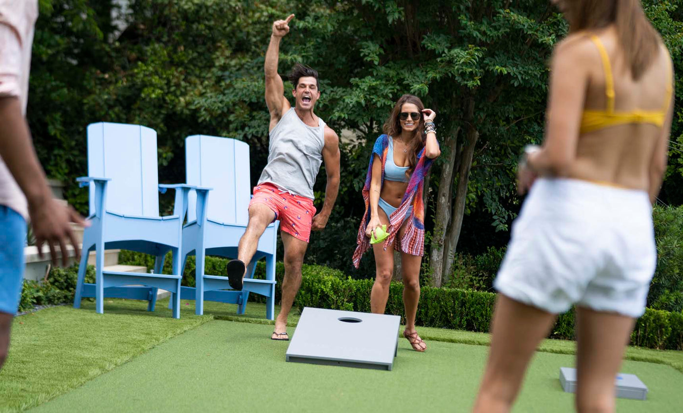Two people enjoy a lively game of Ledge Lounger Cornhole outdoors. One person celebrates while the other smiles with a drink. Nearby, two others stand by the cornhole set, surrounded by large blue chairs and lush greenery in the background.