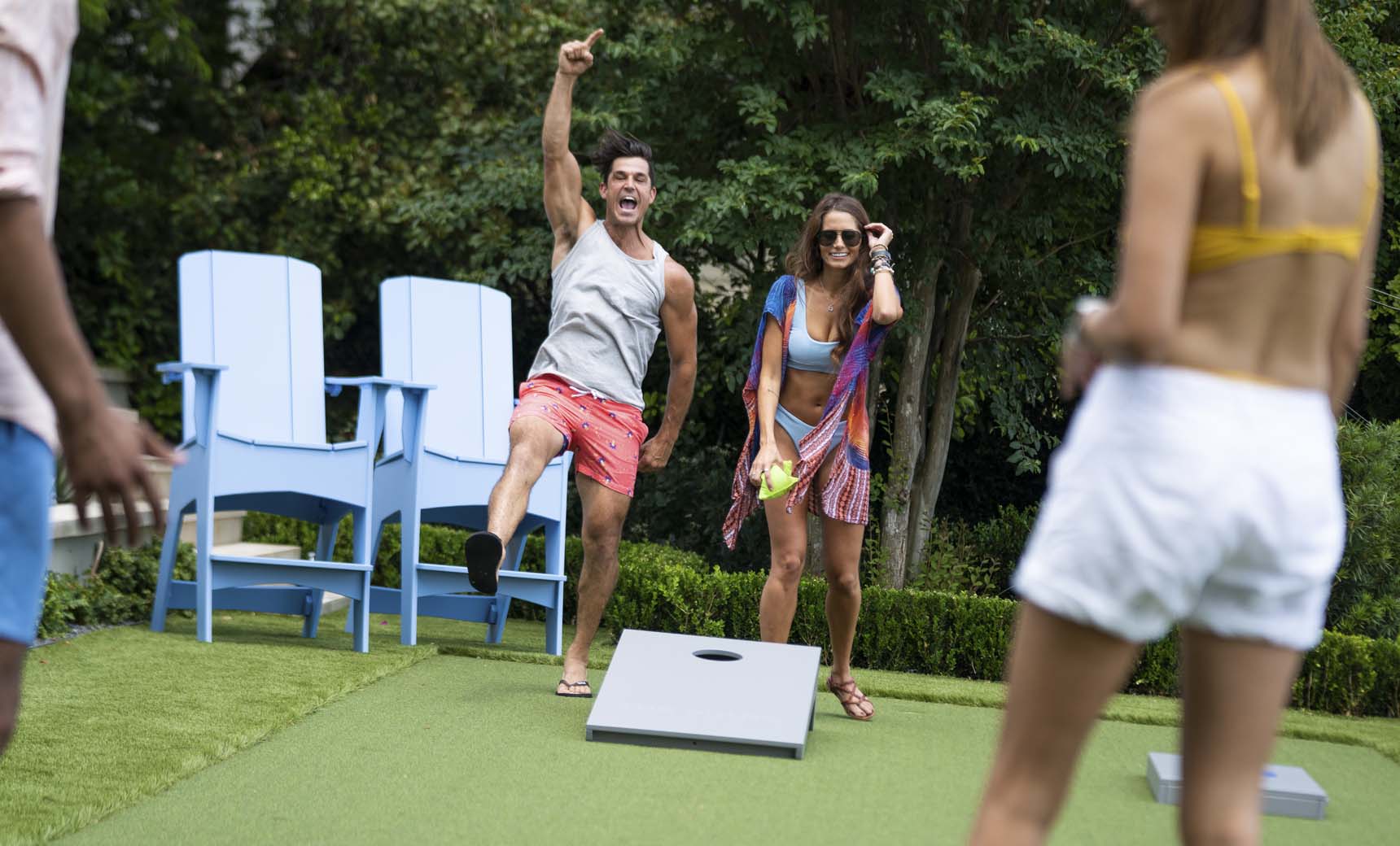 Two people enjoy a lively game of Ledge Lounger Cornhole outdoors. One person celebrates while the other smiles with a drink. Nearby, two others stand by the cornhole set, surrounded by large blue chairs and lush greenery in the background.